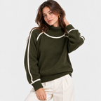 A woman wearing an olive green mock-neck knit sweater with white contrast trim along the shoulders and sleeves, paired with off-white pants, against a plain light gray studio background.