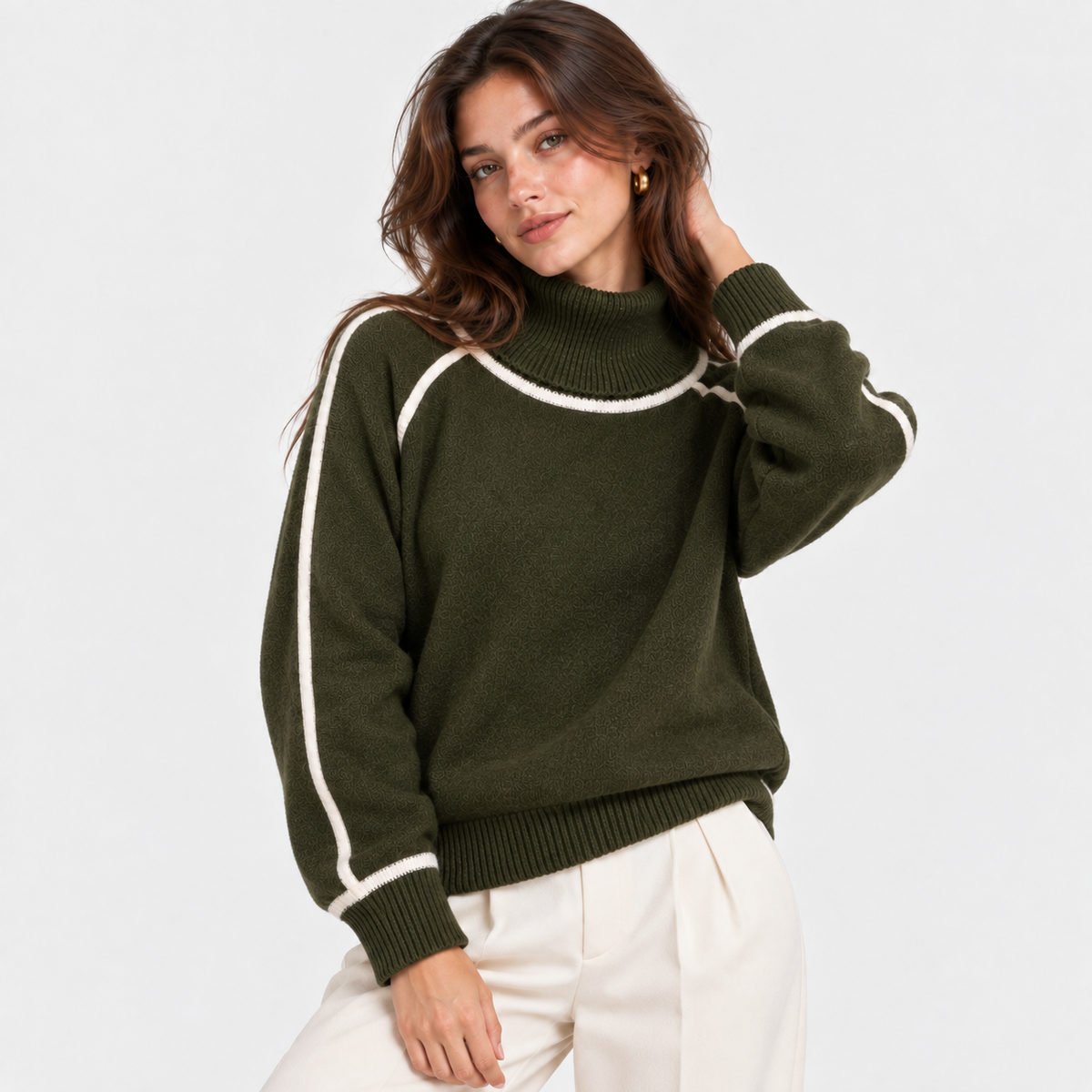 A woman wearing an olive green mock-neck knit sweater with white contrast trim along the shoulders and sleeves, paired with off-white pants, against a plain light gray studio background.