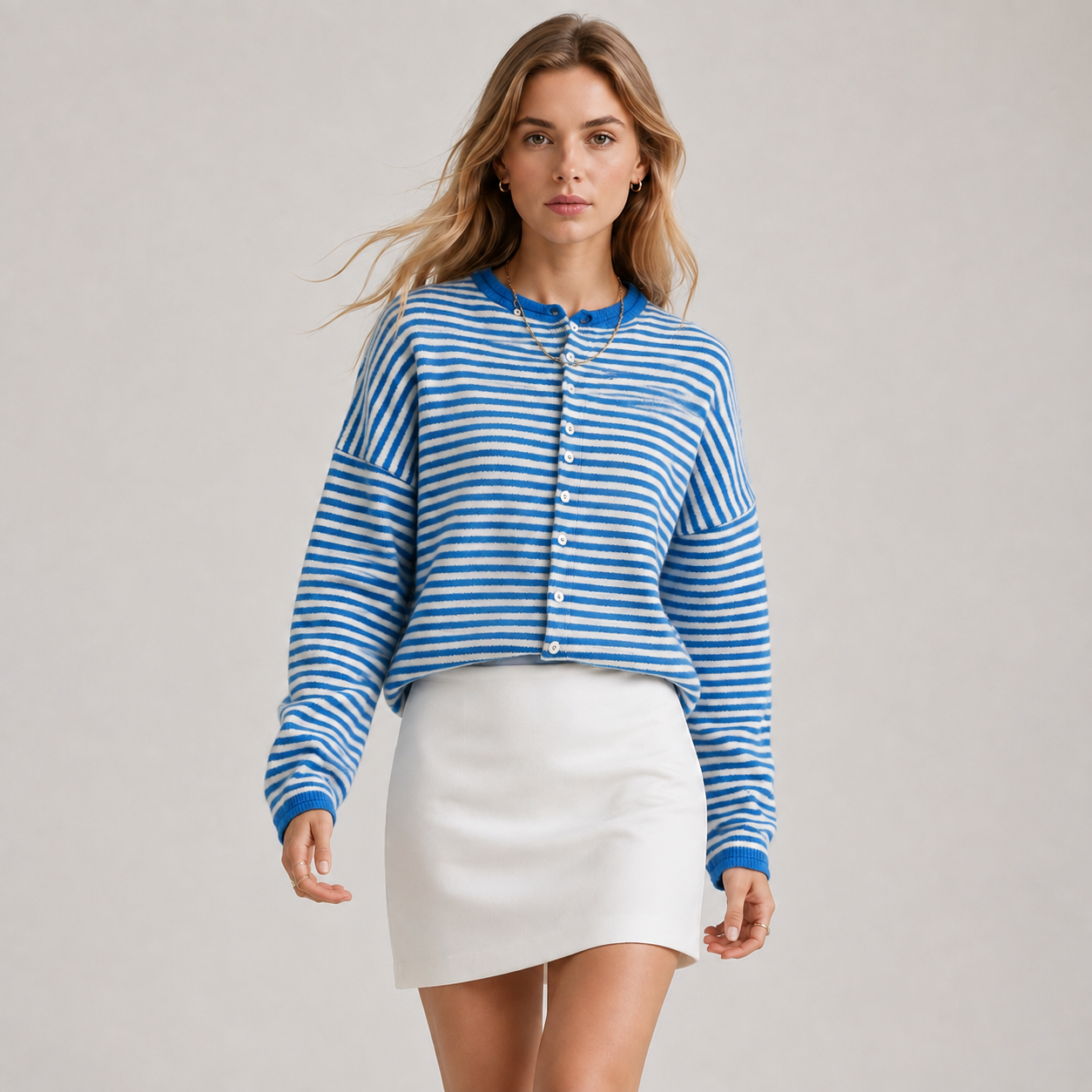 A woman modeling a blue and white striped cropped button-up cardigan sweater paired with a white mini skirt, set against a plain light gray studio background.