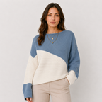 A woman wearing a color-block knit sweater with a dusty blue upper section and sleeves, and an off-white ribbed lower section and sleeves, paired with beige pants against a light gray studio background.