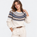 A woman modeling a cream-colored knit sweater with a colorful geometric Fair Isle pattern across the chest and shoulders, paired with cream trousers and a brown belt, against a plain white background.