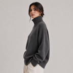 A woman wearing an oversized dark gray ribbed turtleneck sweater, paired with cream-colored trousers, photographed against a neutral light gray studio background.