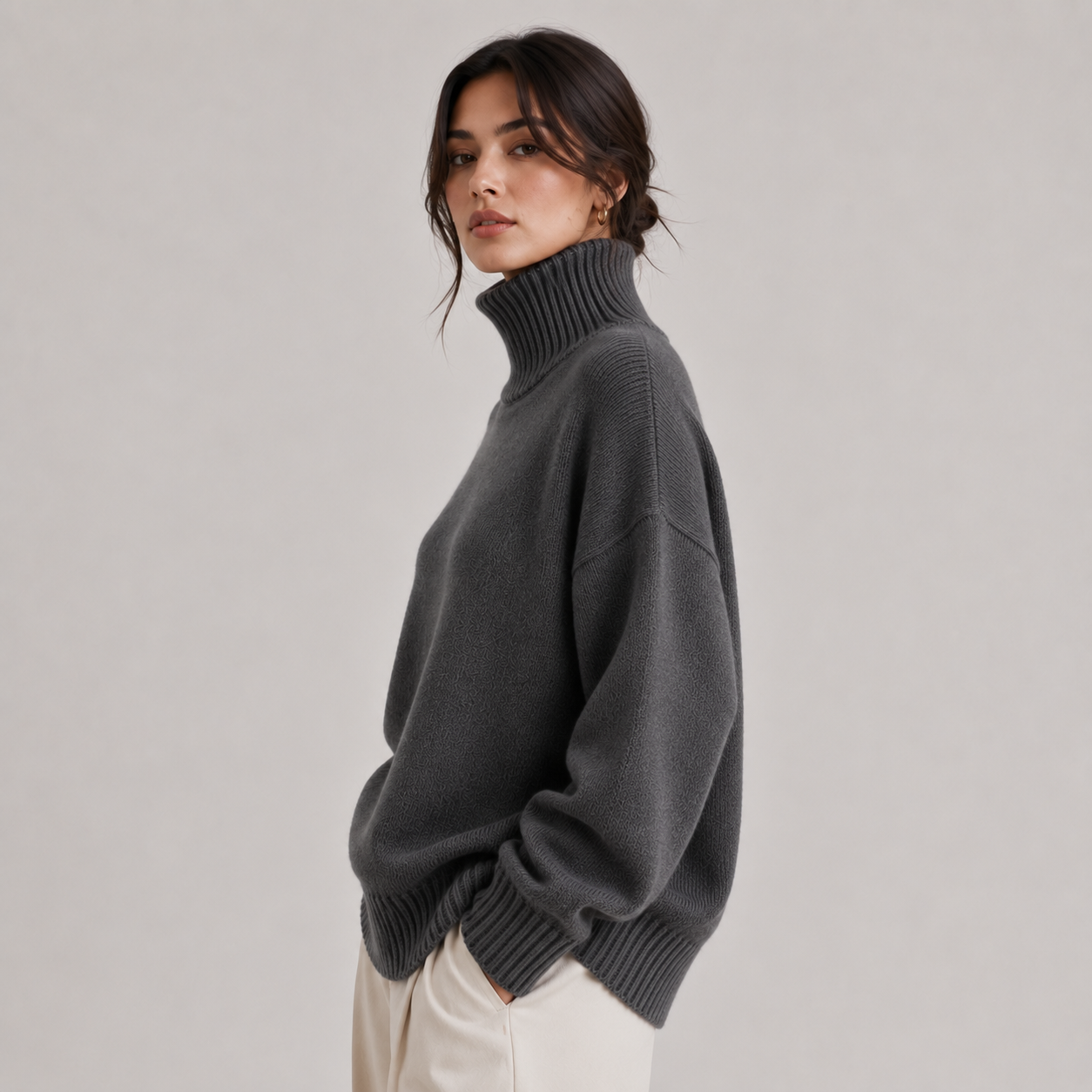 A woman wearing an oversized dark gray ribbed turtleneck sweater, paired with cream-colored trousers, photographed against a neutral light gray studio background.