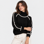 A woman wearing a black mock-neck knit sweater with contrasting white piping details on the shoulders and sleeves, paired with off-white trousers, against a plain white studio background.