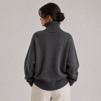A woman shown from the back wearing an oversized dark gray turtleneck sweater and cream-colored pants, set against a plain light gray studio background.