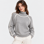 A woman wearing a light gray knit turtleneck sweater with contrasting white ribbed trim along the shoulders, collar, and cuffs, paired with cream-colored trousers, set against a plain light gray background.