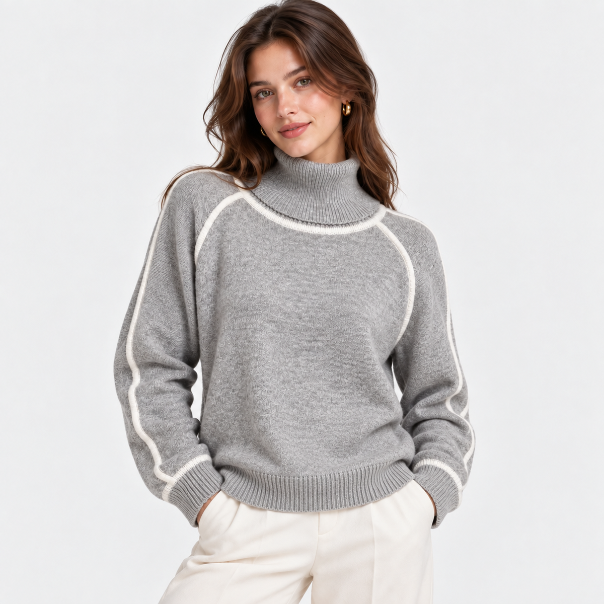 A woman wearing a light gray knit turtleneck sweater with contrasting white ribbed trim along the shoulders, collar, and cuffs, paired with cream-colored trousers, set against a plain light gray background.