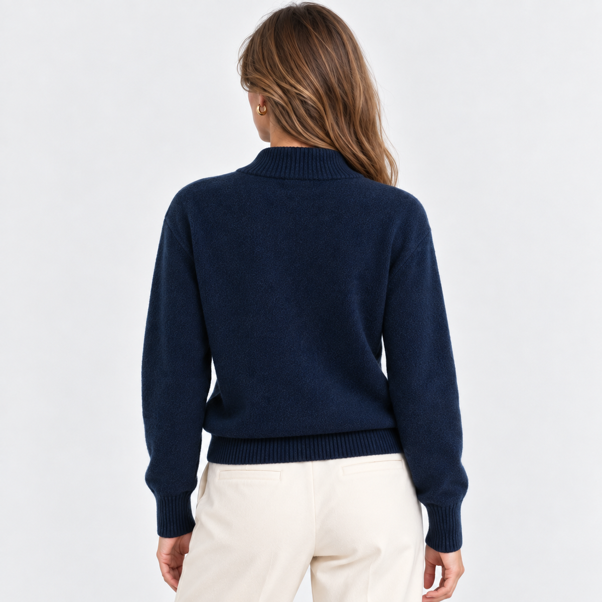 A woman viewed from the back wearing a navy blue knit sweater with a mock neck and cream-colored pants, set against a plain white studio background.