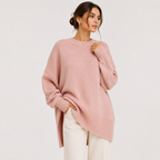 A woman wearing an oversized dusty pink knit sweater with a crew neck and ribbed cuffs, paired with white pants, photographed against a plain light gray studio background.