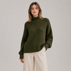 A woman wearing an olive green textured knit mock-neck sweater and cream wide-leg trousers, standing against a neutral light gray studio background.