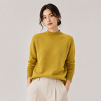 A woman wearing a mustard yellow knit mock neck sweater and light beige trousers, standing against a neutral light gray studio background.