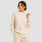 A woman wearing an oversized cream-colored knit sweater and matching trousers, photographed against a plain light gray studio background.