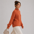 A woman wearing a chunky knit orange mock neck sweater and white trousers, holding a light beige handbag against a plain light gray studio background.