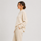A woman wearing an oversized cream knit sweater and matching cream trousers, shown in side profile against a plain light gray studio background.