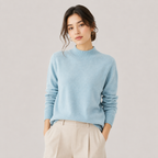 A woman wearing a light blue mock-neck knit sweater and beige trousers, standing against a plain light gray studio background.
