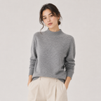 A woman wearing a light gray knit mock neck sweater and cream-colored pleated trousers, standing against a light gray studio background.