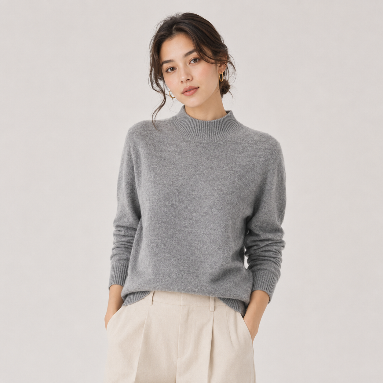 A woman wearing a light gray knit mock neck sweater and cream-colored pleated trousers, standing against a light gray studio background.