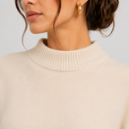 Close-up of a woman wearing a cream ribbed knit mock neck sweater and a small gold hoop earring against a light gray background.