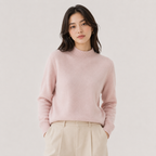 A woman wearing a light pink knit mock-neck sweater and beige trousers, standing against a plain light gray studio background.