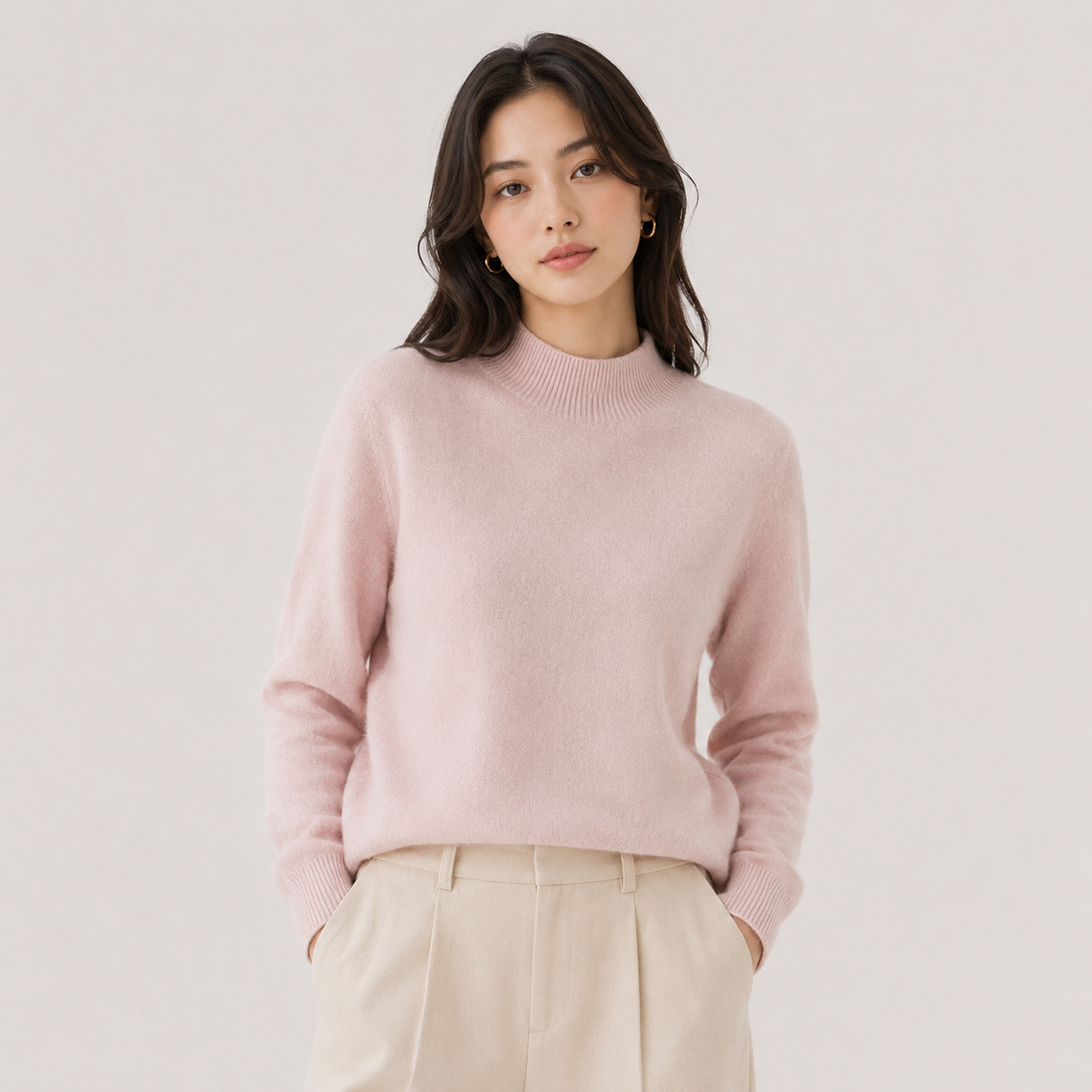A woman wearing a light pink knit mock-neck sweater and beige trousers, standing against a plain light gray studio background.