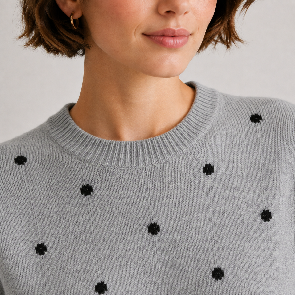 Close-up of a woman wearing a light gray knit sweater with small black embroidered flower-like accents, cropped from the mouth to the chest against a plain light background.