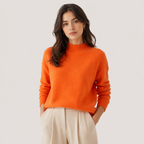 A woman wearing a bright orange knit mock-neck sweater and cream-colored trousers, standing against a light gray studio background.