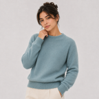 A woman wearing a light blue crewneck knit sweater and cream trousers, posing against a neutral light gray studio background.