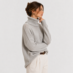A woman wearing a light gray chunky knit turtleneck sweater and cream trousers, shown from the side against a plain light gray studio background.