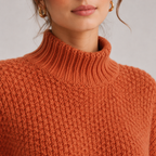 Close-up of a person wearing a chunky knit turtleneck sweater in a warm rust orange color, with gold hoop earrings against a light gray background.