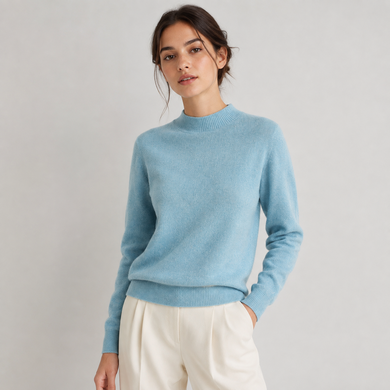 A woman wearing a light blue knit mock-neck sweater and cream-colored trousers, standing against a neutral light gray studio background.