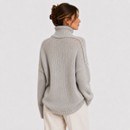 A woman wearing a light gray chunky knit turtleneck sweater, shown from the back and side against a plain light gray studio background.