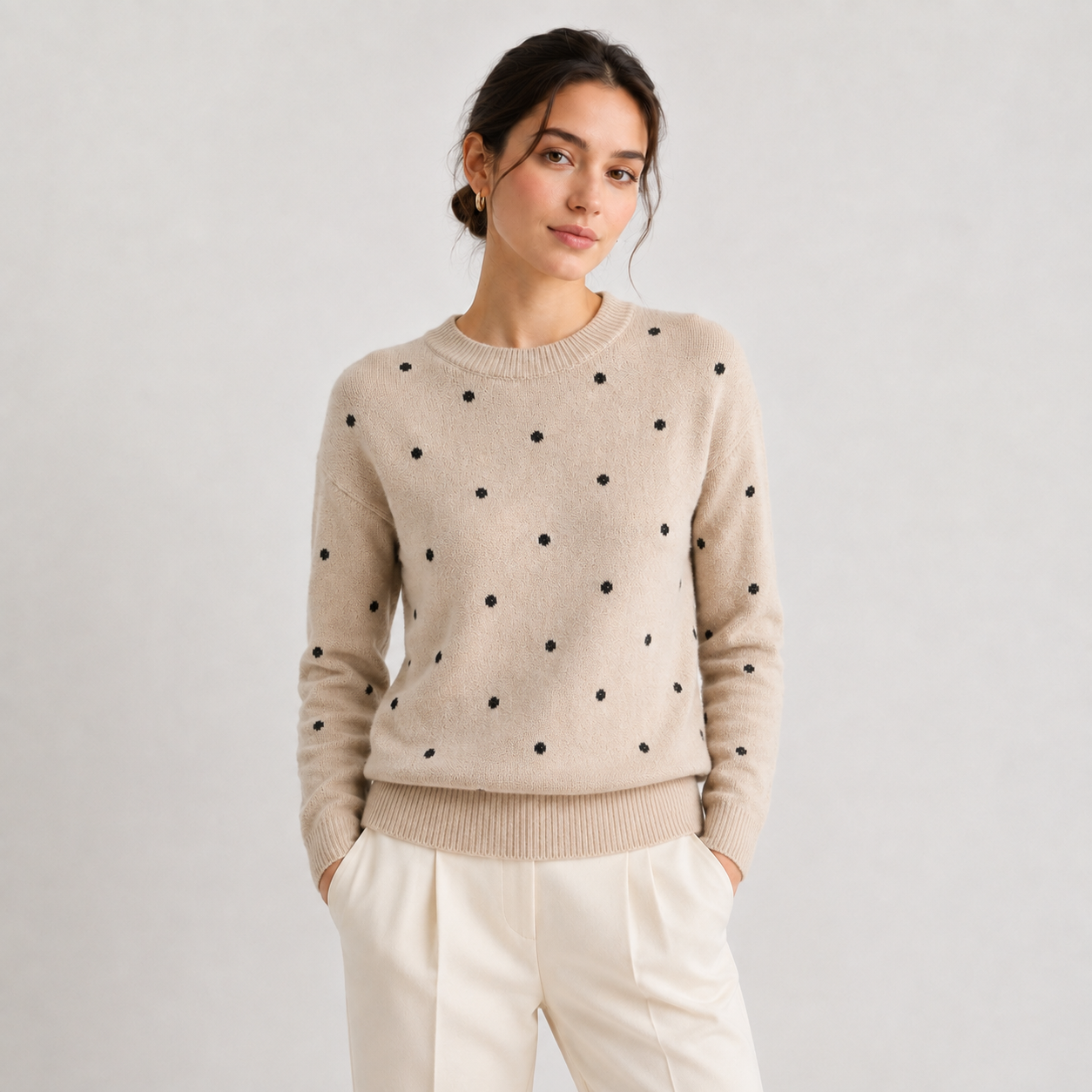 A woman wearing a beige crewneck sweater with black polka dots and cream-colored trousers, standing against a light gray studio background.