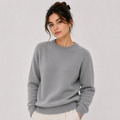 A woman wearing a light gray crewneck knit sweater and off-white trousers, standing against a plain light gray studio background.