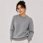 A woman wearing a light gray crewneck knit sweater and off-white trousers, standing against a plain light gray studio background.