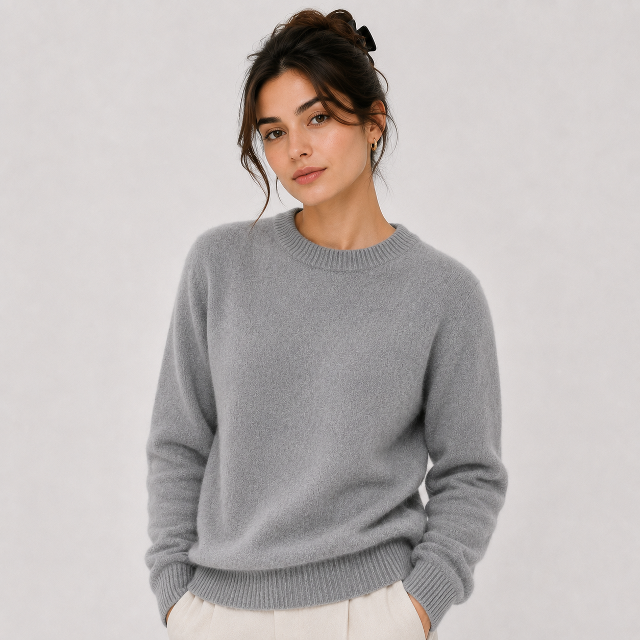 A woman wearing a light gray crewneck knit sweater and off-white trousers, standing against a plain light gray studio background.