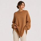 A woman wearing an oversized camel brown knit sweater and cream pants, standing against a light gray studio background.