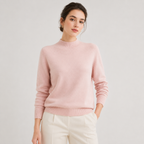 A woman wearing a light pink mock neck knit sweater and cream-colored trousers, standing against a neutral light gray studio background.