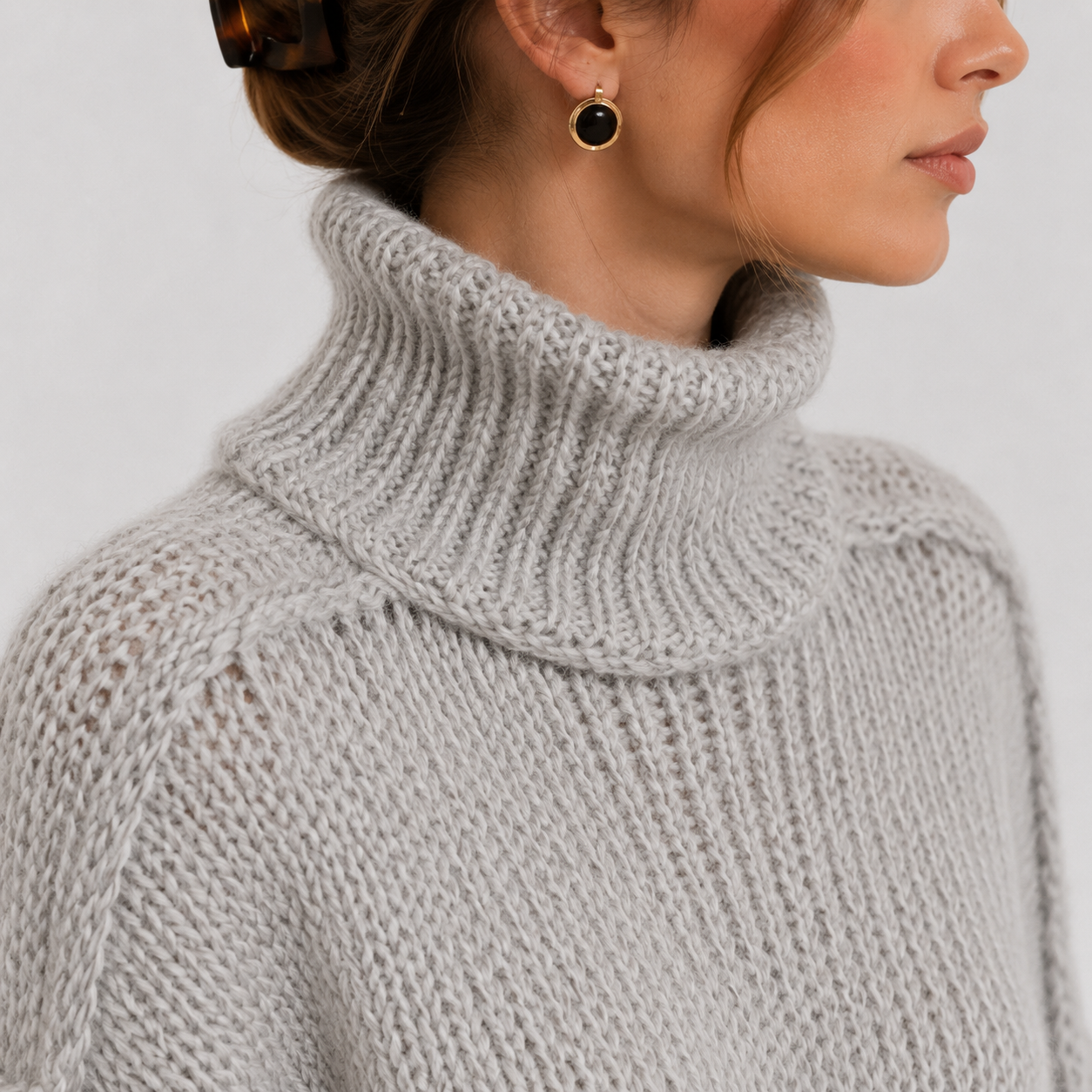 Close-up of a woman wearing a light gray chunky knit turtleneck sweater, with a small gold hoop earring featuring a dark round stone visible.