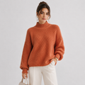 A woman wearing a chunky knit mock neck sweater in a warm rust orange color, paired with white trousers, standing against a light gray studio background.