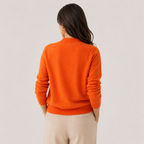 A woman viewed from the back wearing a bright orange knit sweater and light beige trousers against a plain light gray studio background.