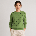 A woman wearing a sage green crewneck sweater with black polka dots, paired with light beige trousers, standing against a light gray studio background.