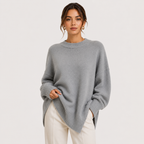 A woman wearing an oversized light gray knit sweater and cream pants, standing against a plain light gray studio background.