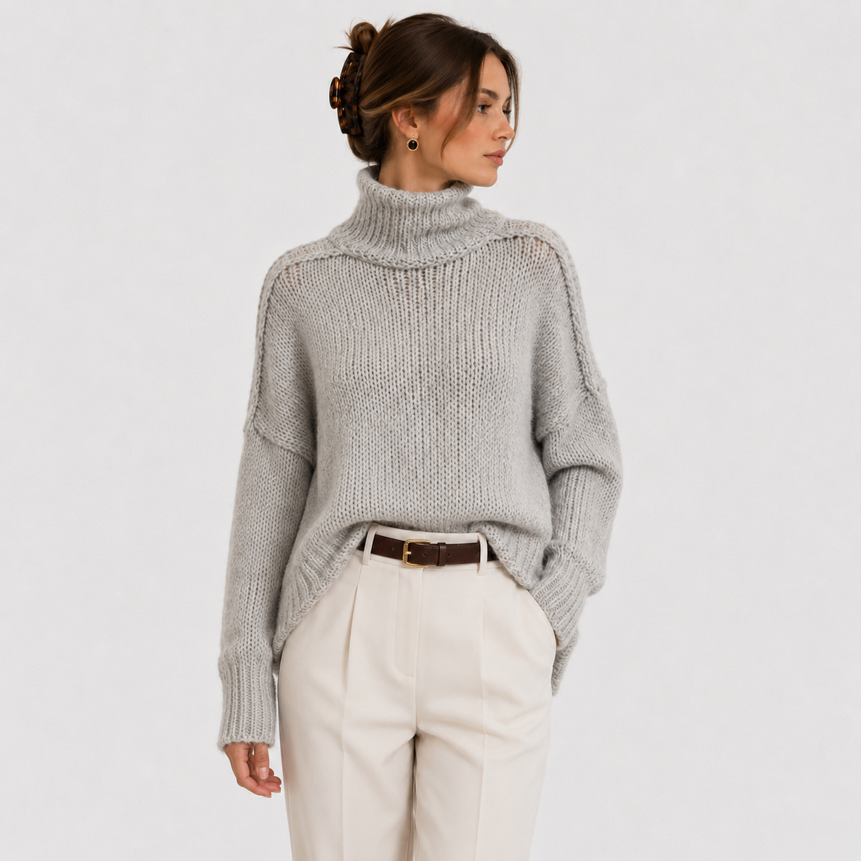 A woman wearing a light gray chunky knit turtleneck sweater and cream trousers, photographed against a plain light gray studio background.