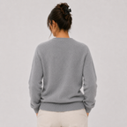 A woman viewed from behind wearing a light gray knit crewneck sweater and cream-colored pants, set against a plain light gray studio background.