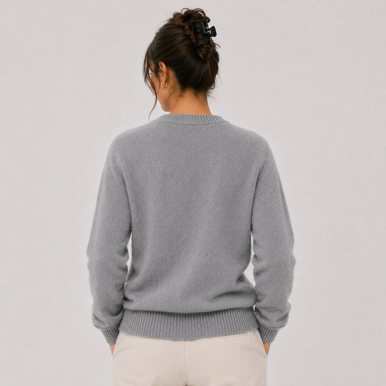 A woman viewed from behind wearing a light gray knit crewneck sweater and cream-colored pants, set against a plain light gray studio background.