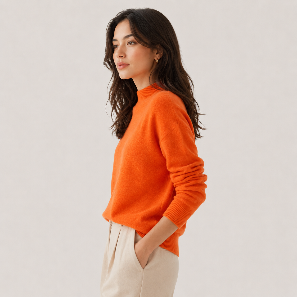 A woman wearing a bright orange knit mock-neck sweater and cream trousers, shown in profile against a light gray studio background.
