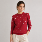 A woman wearing a red crewneck sweater with white polka dots, paired with cream trousers, standing against a light gray studio background.
