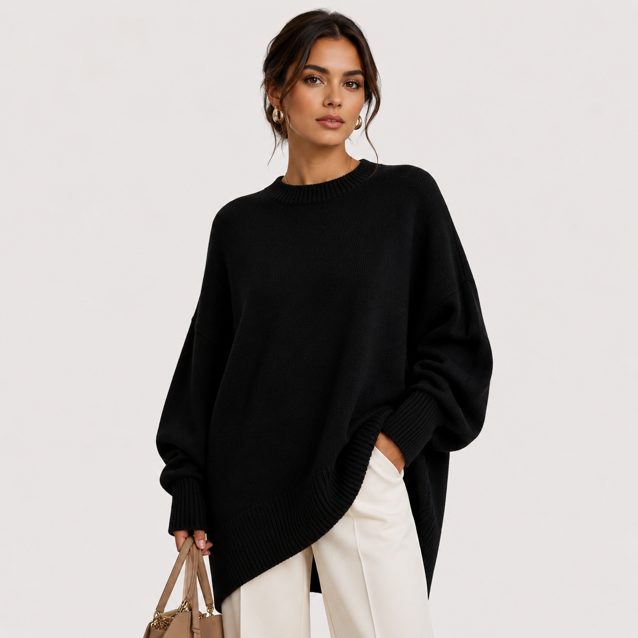 A woman modeling an oversized black knit sweater with a crew neck and relaxed fit, paired with off-white trousers and holding a beige handbag against a plain light gray studio background.