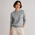A woman wearing a light gray crewneck sweater with black polka dots, paired with light beige trousers, photographed against a neutral light gray studio background.