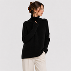 A woman wearing a black ribbed knit turtleneck sweater and cream trousers, photographed against a plain light gray studio background.